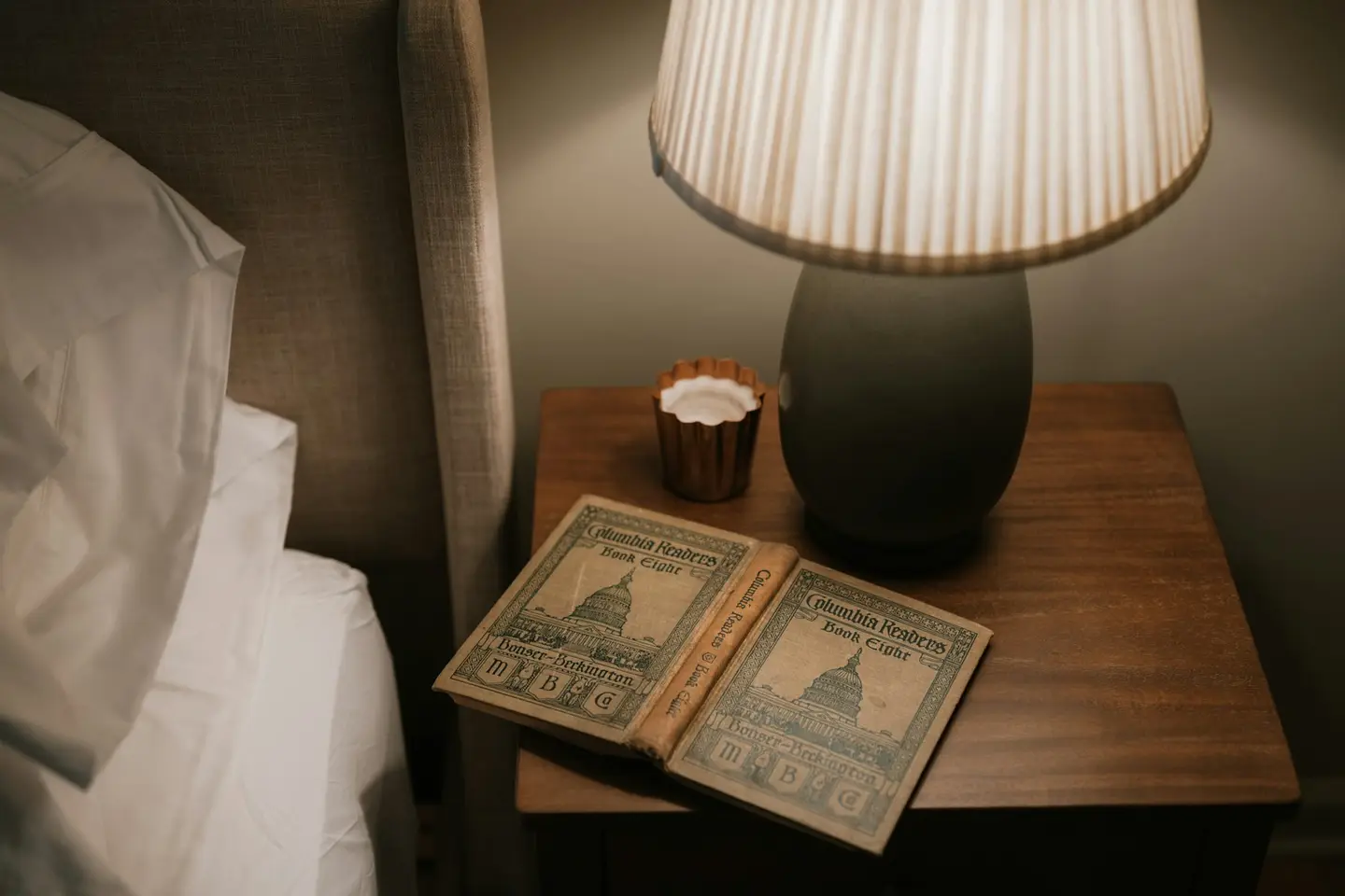 An open book lies on a bedside table under a lit lamp, next to a bed.